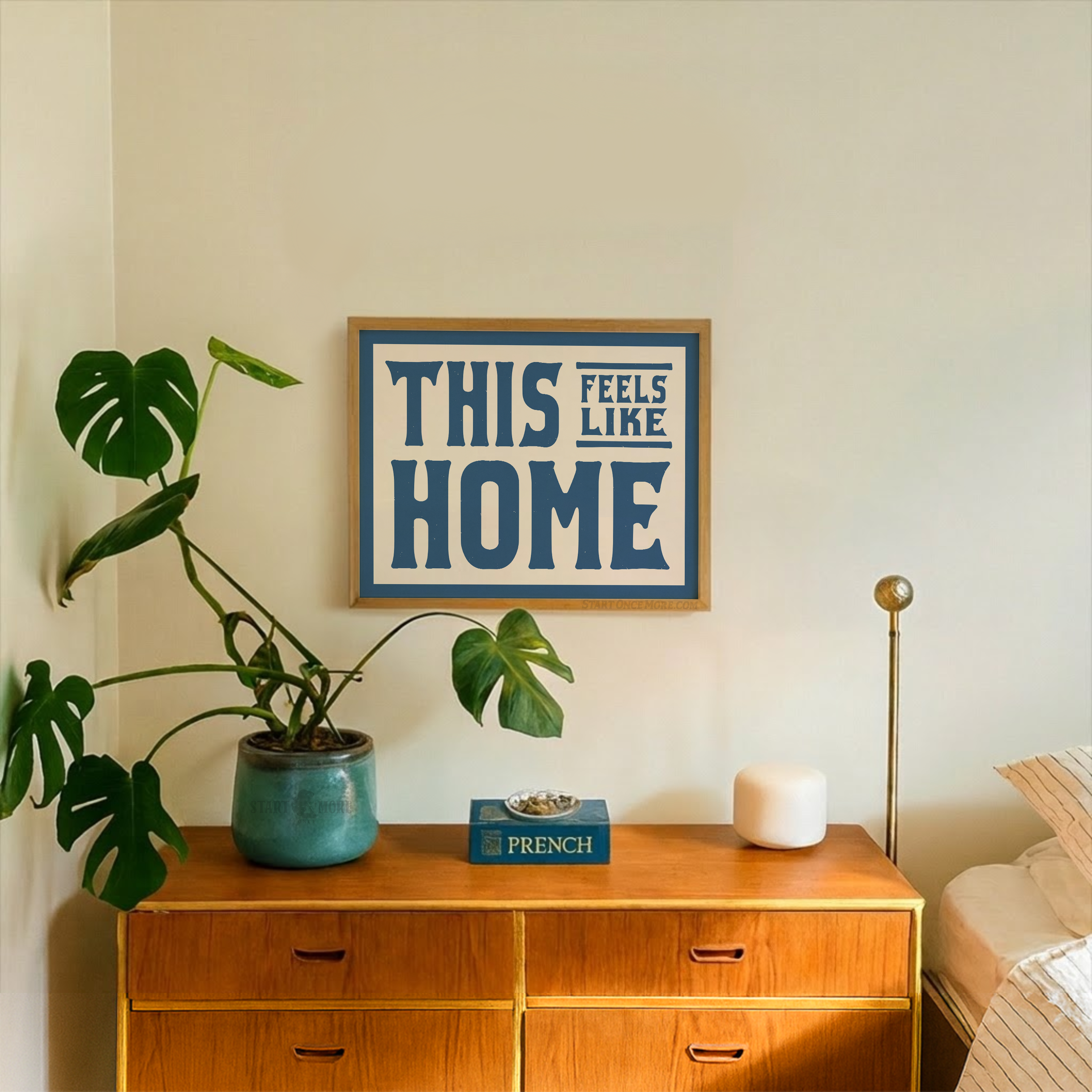 Wooden dresser with a plant, book, and candle, framed 'This Feels Like Home' sign on the wall.